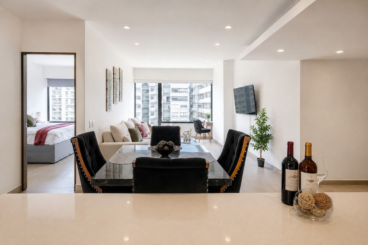 A modern living room with a dining area, a sofa set, a TV mounted on the wall, and a view of the cityscape through large windows.