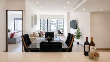 A modern living room with a dining area, a sofa set, a TV mounted on the wall, and a view of the cityscape through large windows.