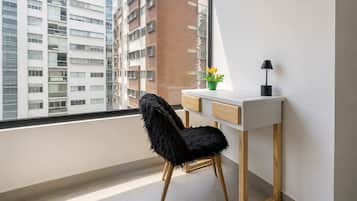 Quarto moderno com vista para edifícios altos, uma escrivaninha branca, uma cadeira preta, uma pequena planta e uma luminária de mesa.