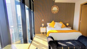 Habitació d'hotel moderna amb un llit gran, una tauleta de nit i vistes a la ciutat a través de finestres de sostre a terra.