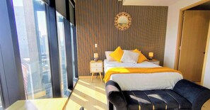 Moderni hotellihuone, jossa on suuri sänky, yöpöytä, sängyn vieressä oleva pöytä ja näkymä kaupunkiin lattiasta kattoon ulottuvien ikkunoiden kautta.
