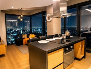 A modern kitchen with a central island, a dining area with a sofa, and a city view through large windows. - 24th Floor CDMX Condo: Pool, Gym, Panoramic Views (Mexico City)