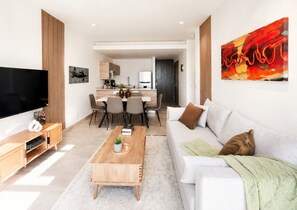 Un salon moderne avec un téléviseur à écran plat, une table basse et un canapé. On aperçoit à l’arrière-plan un coin repas avec des chaises et une table.
