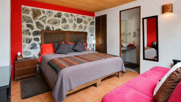 Ein Schlafzimmer mit einer Steinsäule, einem Bett mit farbenfroher Decke, einem Holz-Kopfteil und einer roten Akzentwand.
