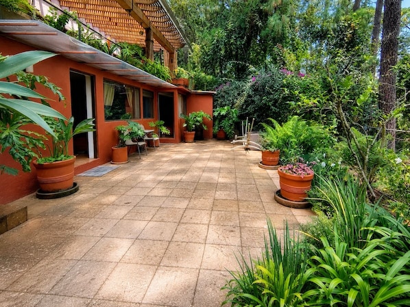 A covered outdoor area with a tiled floor, surrounded by potted plants and trees.
