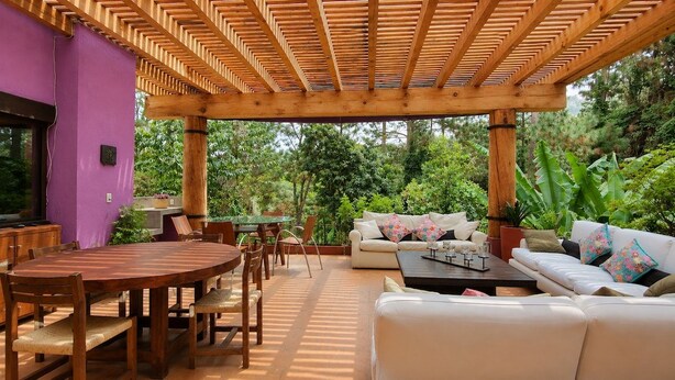 An outdoor patio with a wooden pergola, a round wooden table with chairs, a white sofa with cushions, and a small coffee table. - Swallow | Forest, fireplace and jacuzzi (Valle de Bravo)