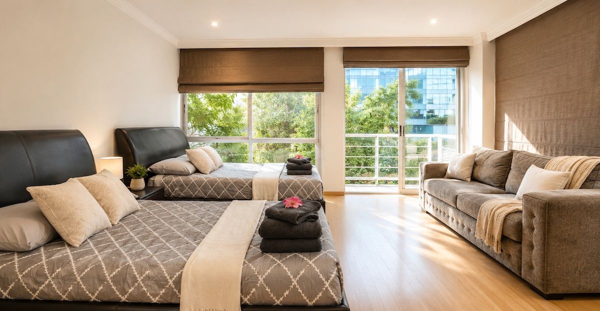 Ein Hotelzimmer mit zwei Betten, einem Sofa und einem großen Fenster mit Blick auf Grünflächen und Gebäude.