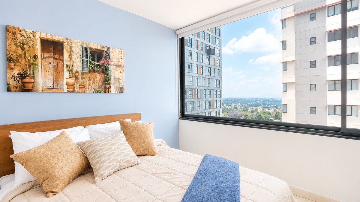 A bedroom with a bed, pillows, a blue throw blanket, a window with a city view, and a wall-mounted artwork.