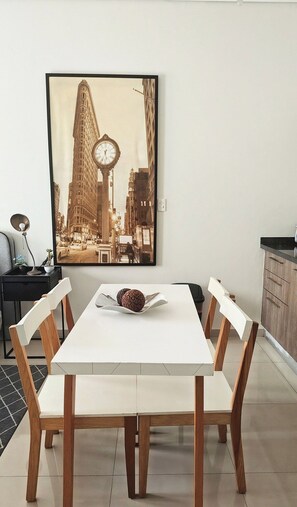 Un coin repas avec une table blanche et des chaises en bois, un tableau représentant un paysage urbain encadré accroché au mur, et une petite plante en pot posée sur la table d’appoint.