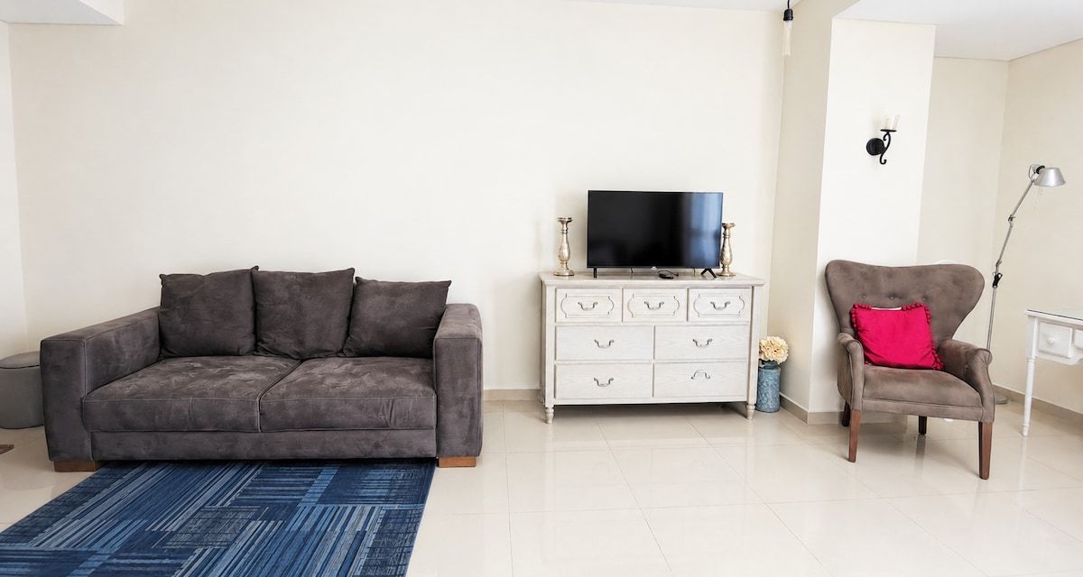 Ein Wohnzimmer mit einer Couch, einem Fernseher auf einem Schrank, einem Sessel, einem weißen Kommoden und einem blauen Teppich.