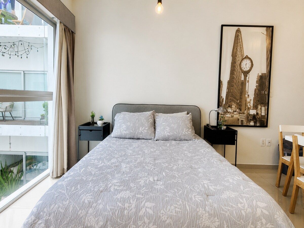 A bedroom with a large bed, a nightstand, a desk, a chair, and a window with curtains.