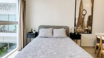 A bedroom with a large bed, a nightstand, a desk, a chair, and a window with curtains.