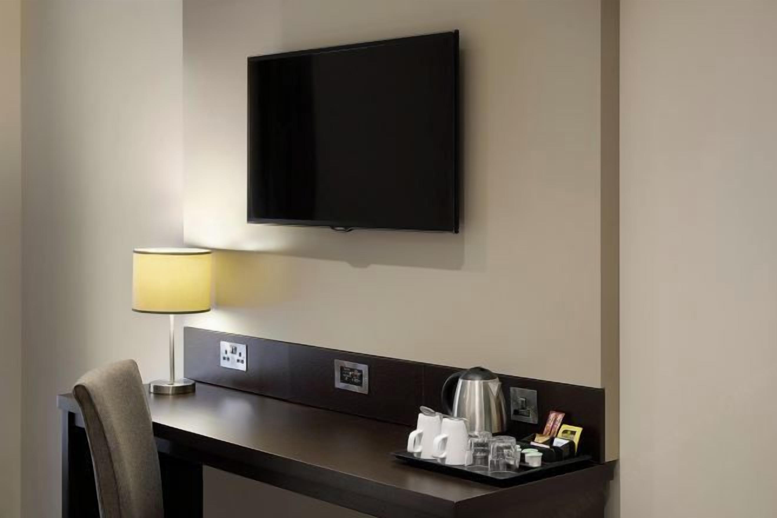 A hotel room with a flat-screen TV mounted on the wall, a desk with a lamp, and a tray with coffee cups and a coffee pot.