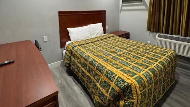 A hotel room with a bed, a wooden desk, a nightstand, a wall-mounted air conditioner, and a window with curtains. - Moonlight Inn (Gardena)