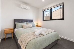 A bedroom with a bed, a nightstand, a bedside lamp, and a window. - Longfin Cove - Villa 1 by Ready Set Stay (San Remo)