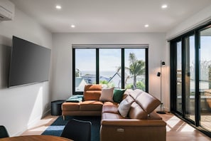 A modern living room with a sectional sofa, a flat-screen TV, and a large window offering a view of the outdoors. - Longfin Cove - Villa 1 by Ready Set Stay (San Remo)