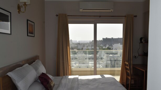A hotel room with a bed, a desk, and a view of the cityscape through a large window. - Flower Valley: Experience Serene Luxury (Sohna)