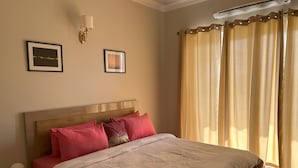 A bedroom with a bed, a nightstand, a wall-mounted lamp, and a window with curtains. - Flower Valley: Experience Serene Luxury (Sohna)