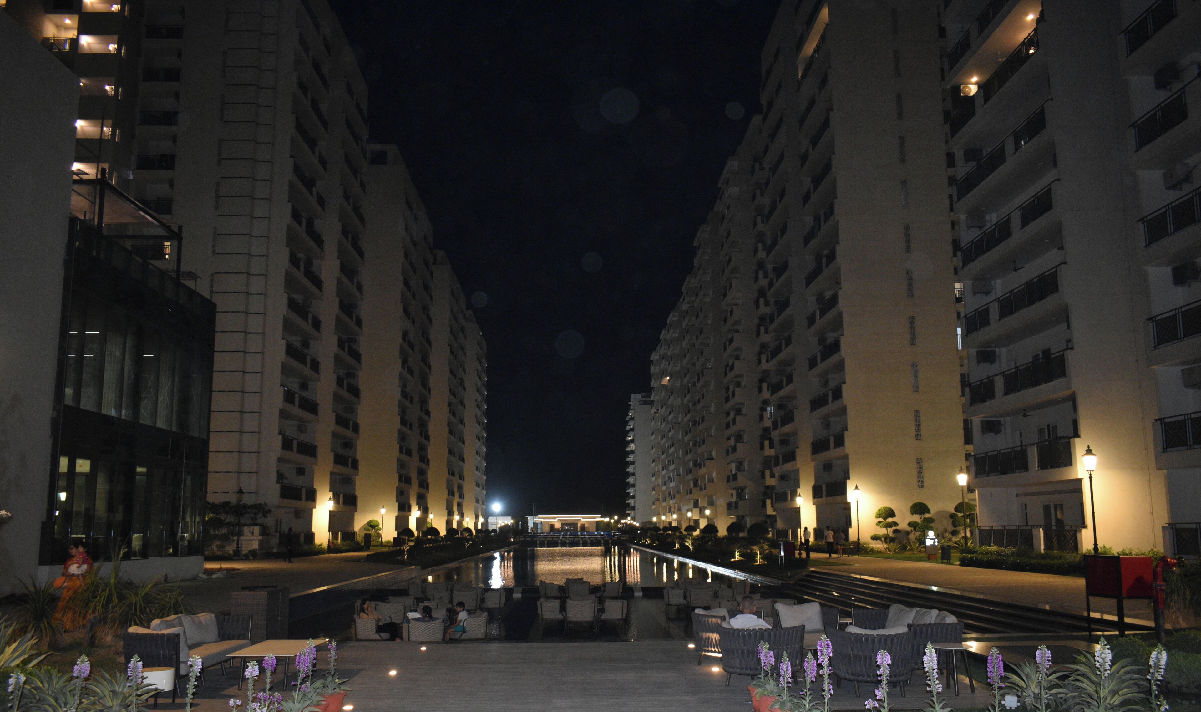 A nighttime view of a hotel corridor with multiple floors, outdoor seating, and a pool area.