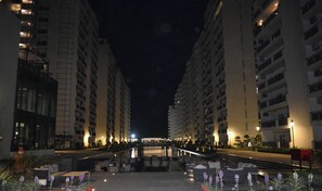 Vista nocturna d'un passadís d'hotel amb diversos pisos, zones d'seients exteriors i una zona de piscina.