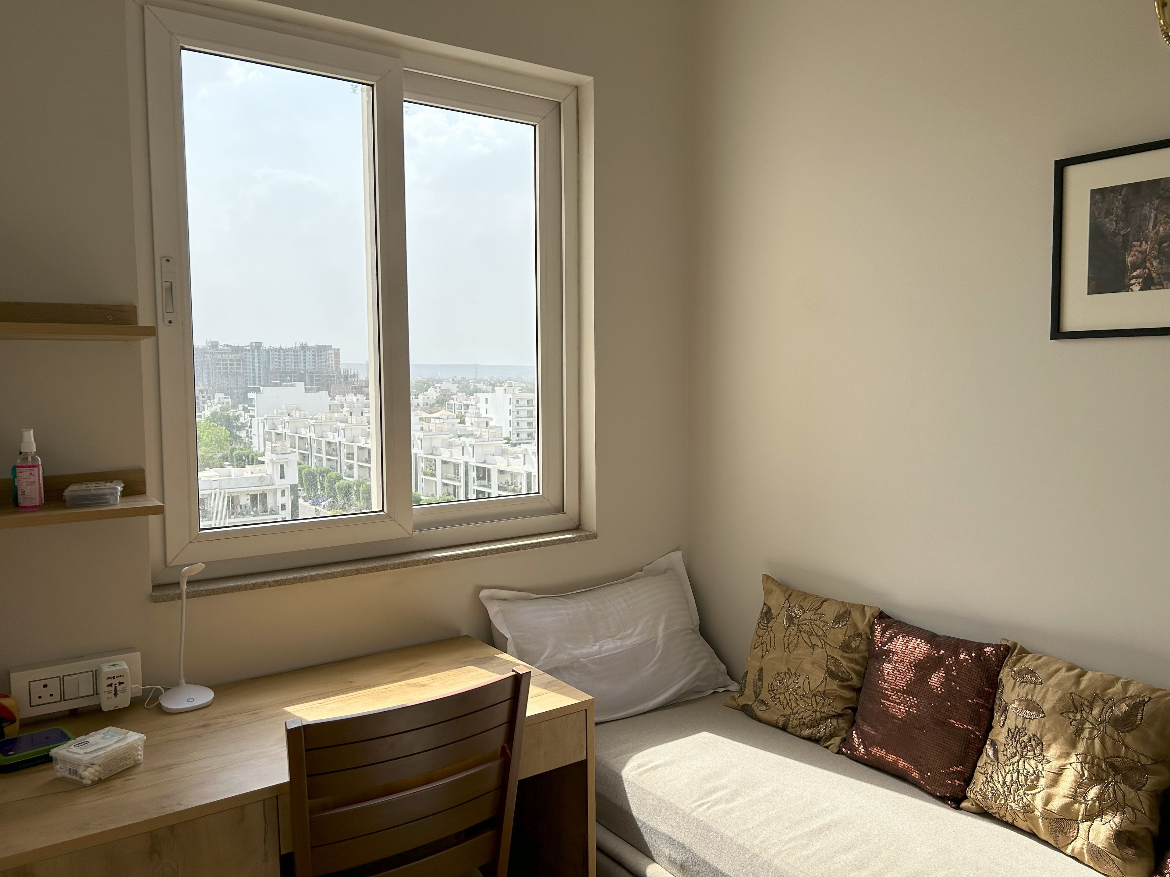A bedroom with a large window, a desk, a chair, a bed with patterned pillows, and a framed picture on the wall.