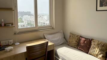 A bedroom with a large window, a desk, a chair, a bed with patterned pillows, and a framed picture on the wall.