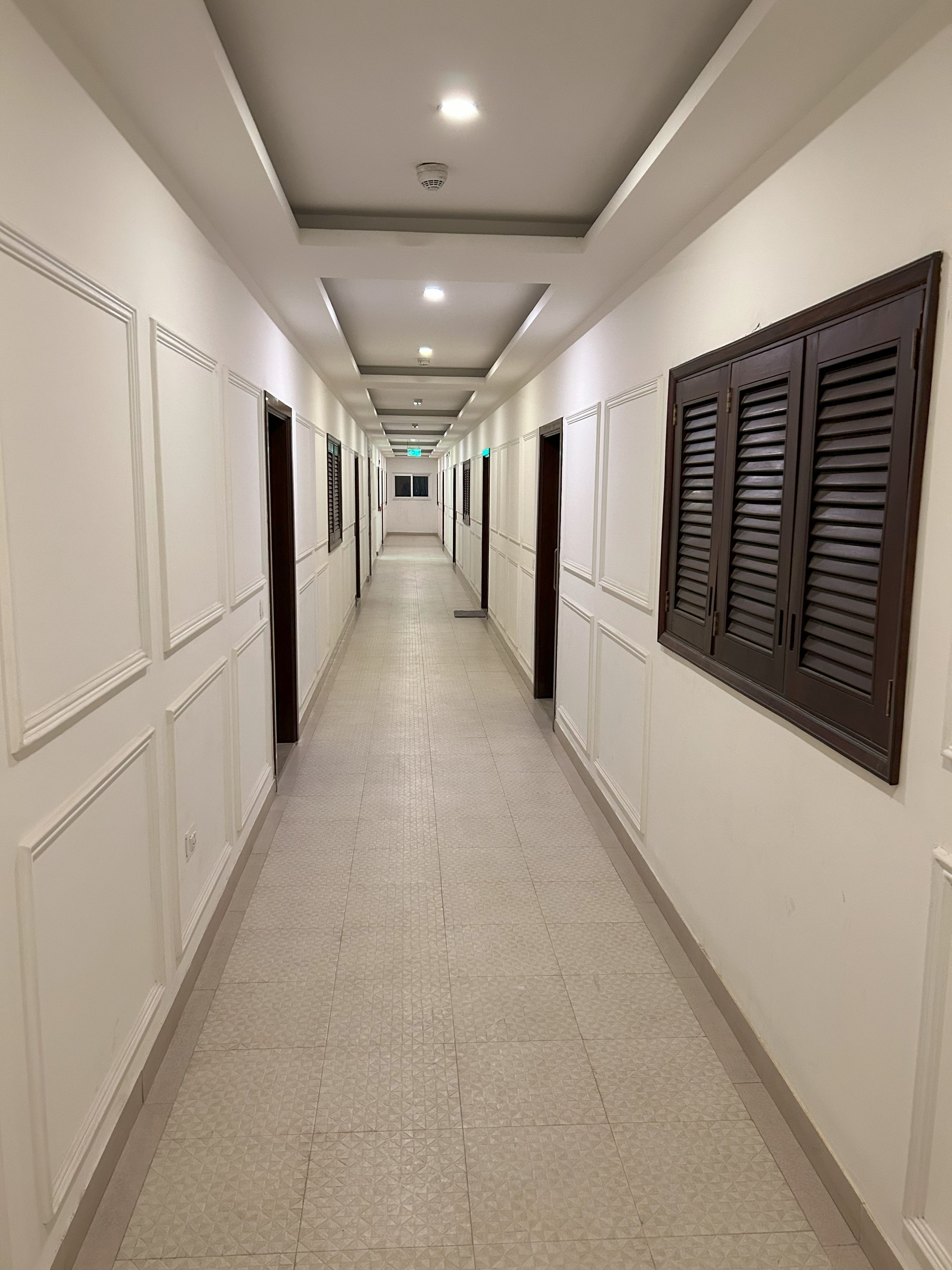 A long, narrow hotel corridor with doors on both sides, a tiled floor, and a ceiling with recessed lighting.