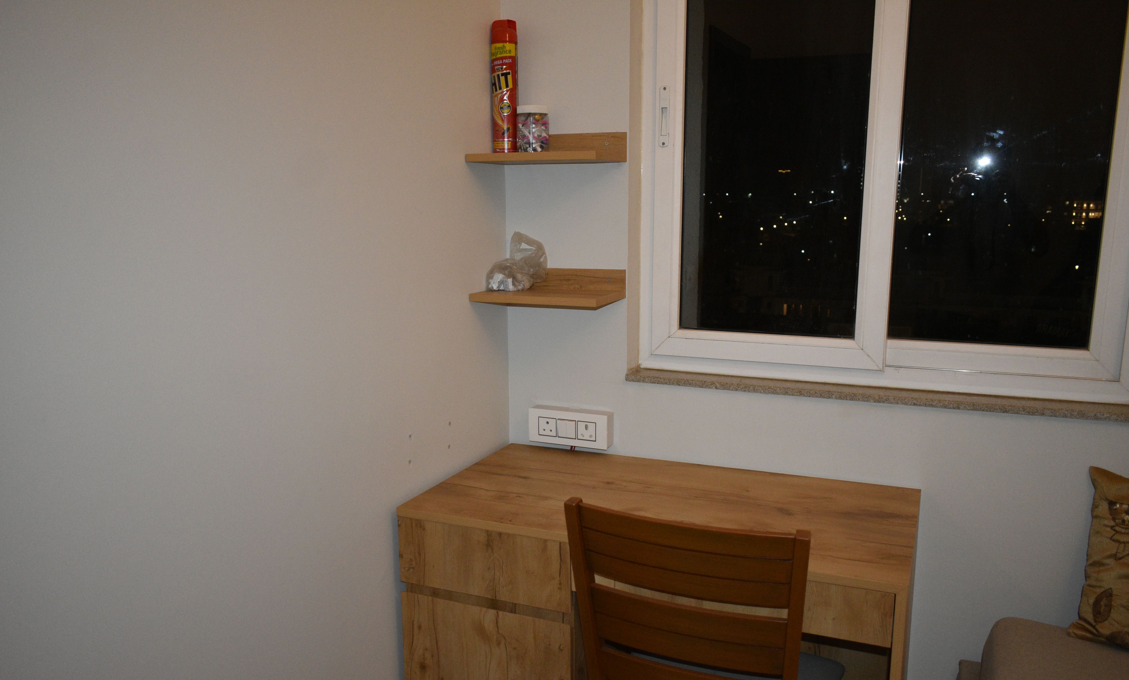 A small room with a wooden desk, a chair, a window with a view of the city at night, and a shelf with a can and a plastic bag.