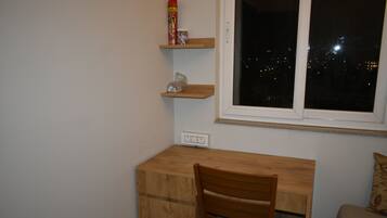 A small room with a wooden desk, a chair, a window with a view of the city at night, and a shelf with a can and a plastic bag.