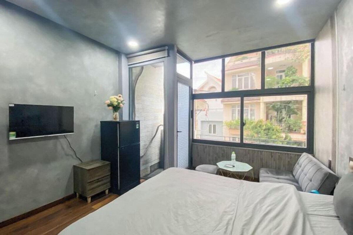 Ein Schlafzimmer mit einem Bett, einem kleinen Tisch, einem Fernseher und einem Fenster mit Blick auf Gebäude.