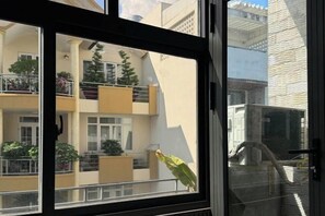 Blick durch ein Fenster auf ein Gebäude mit mehreren Balkonen, von denen einige mit Pflanzen bepflanzt sind, und einem wolkenlosen Himmel.