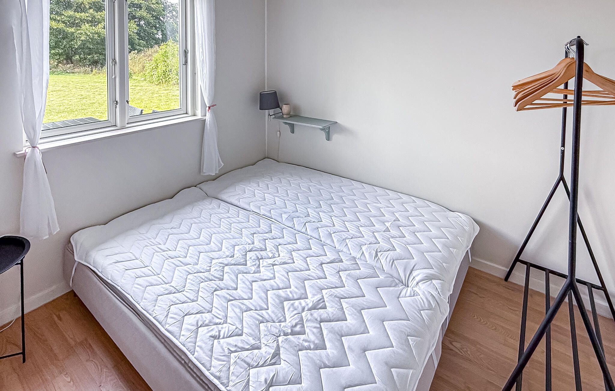 A single bed with a white quilt, a bedside table with a lamp, and a window with curtains.
