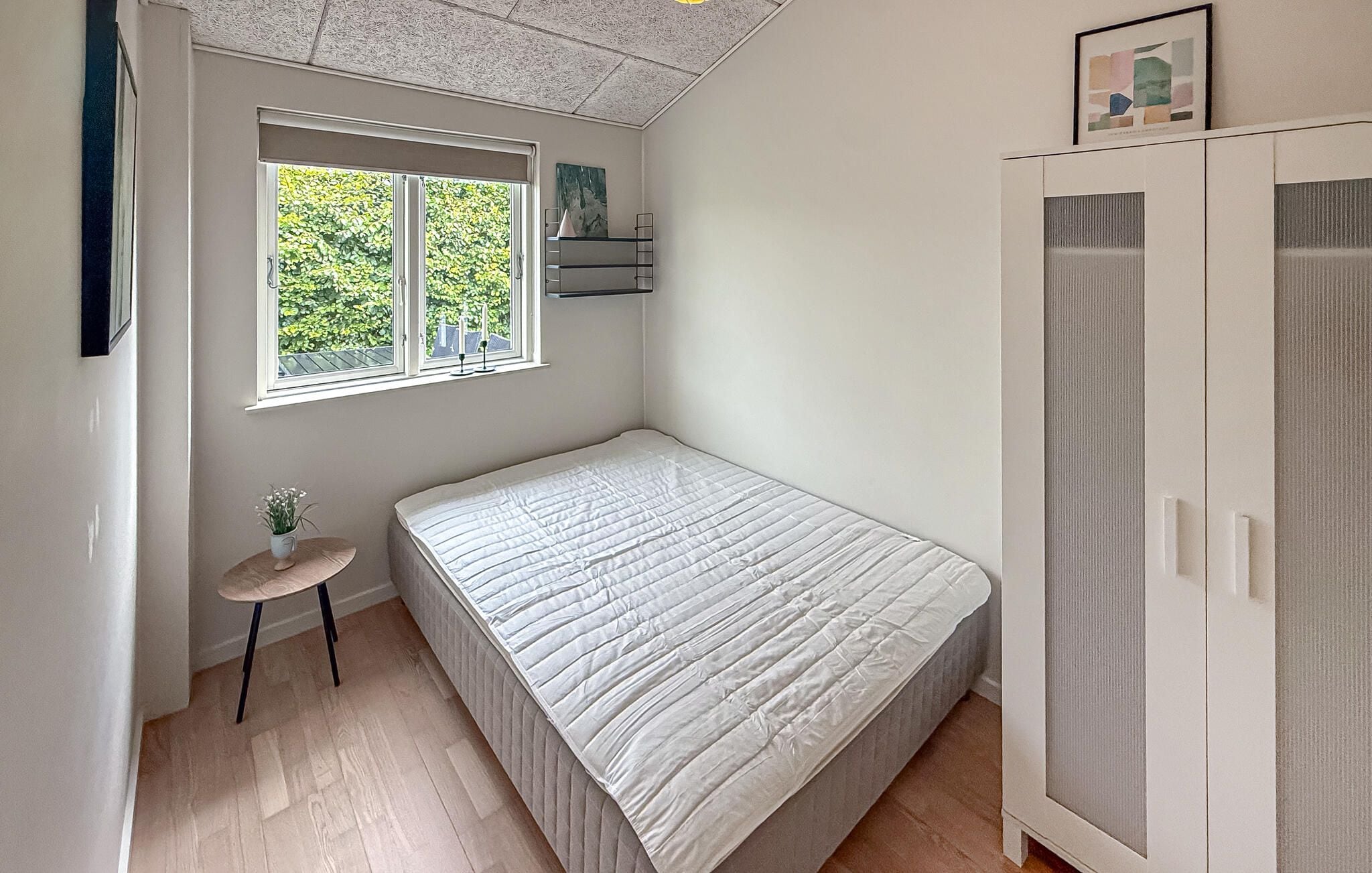 Quarto com uma cama de solteiro, uma mesinha com uma planta, uma janela com vista para o verde e um guarda-roupa.
