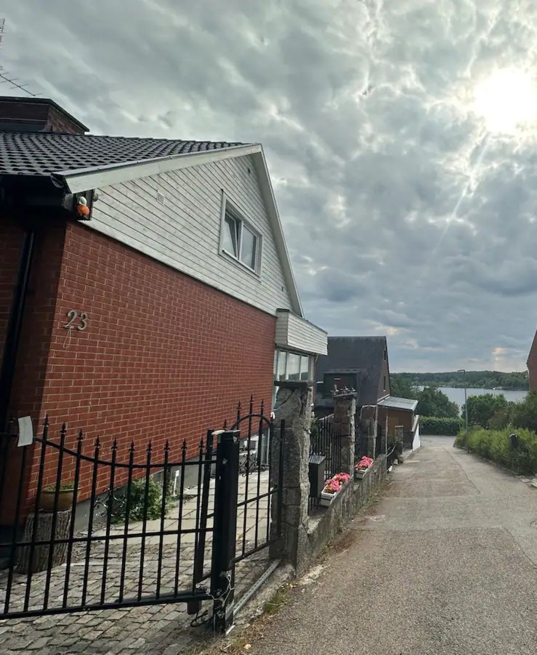 A residential street with a brick house numbered 23, a metal fence, and a paved road leading to a body of water.