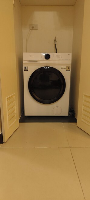 A compact, white front-loading washing machine with a control panel, a 10-year warranty label, and a water inlet hose. - Spacious Family-Friendly 2BR Near BGC & McKinley! (Pasig)