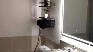 Une salle de bain comprenant des toilettes, un lavabo et un miroir. On y trouve des étagères avec des articles de toilette et un espace douche.