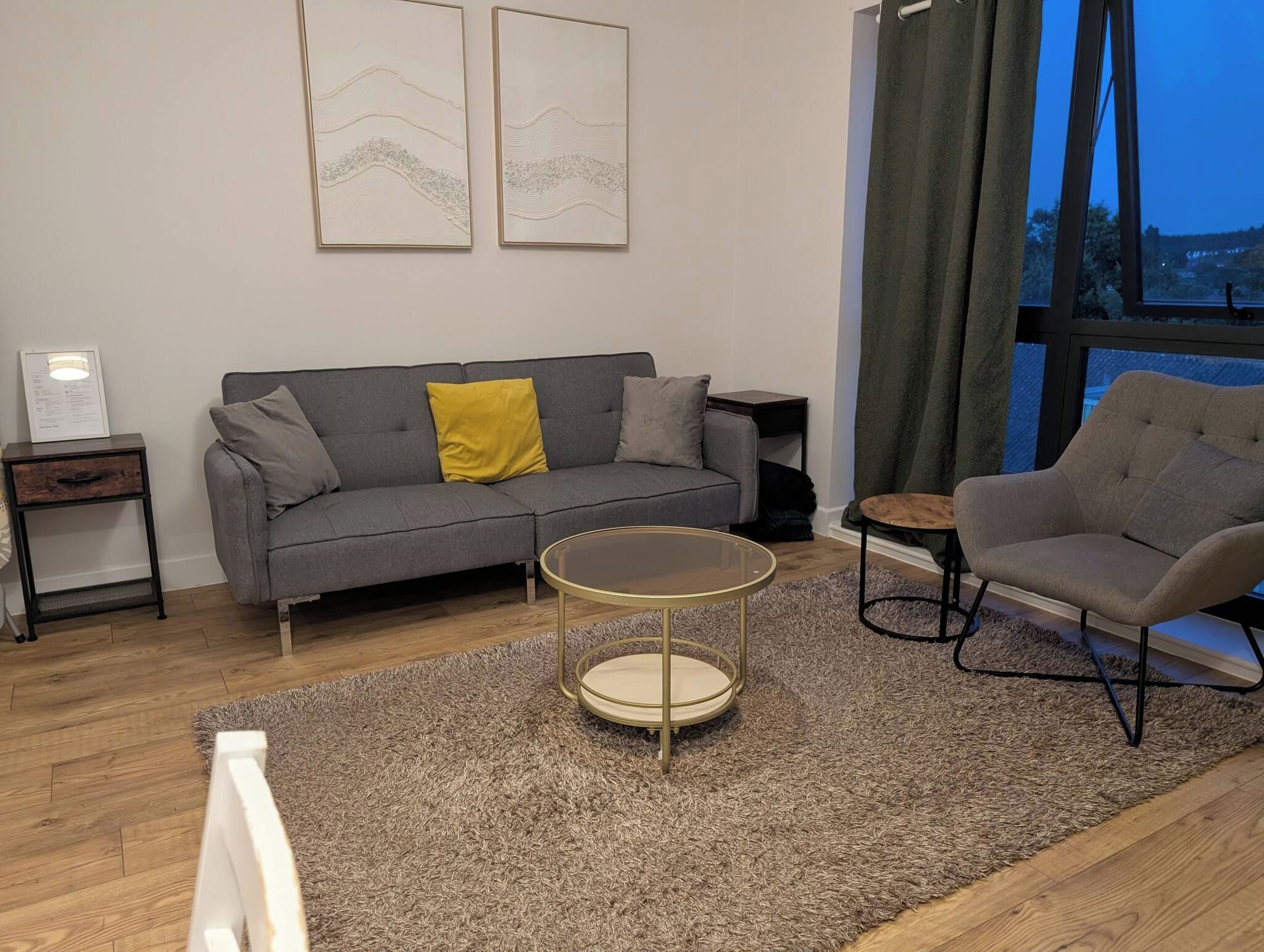 Uma sala de estar moderna com um sofá cinzento, uma mesa de centro redonda e uma poltrona cinzenta. Há duas obras de arte emolduradas na parede e uma janela com cortinas.