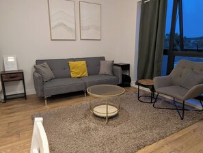 A modern living room with a grey sofa, a round coffee table, and a grey armchair. There are two framed artworks on the wall and a window with curtains.