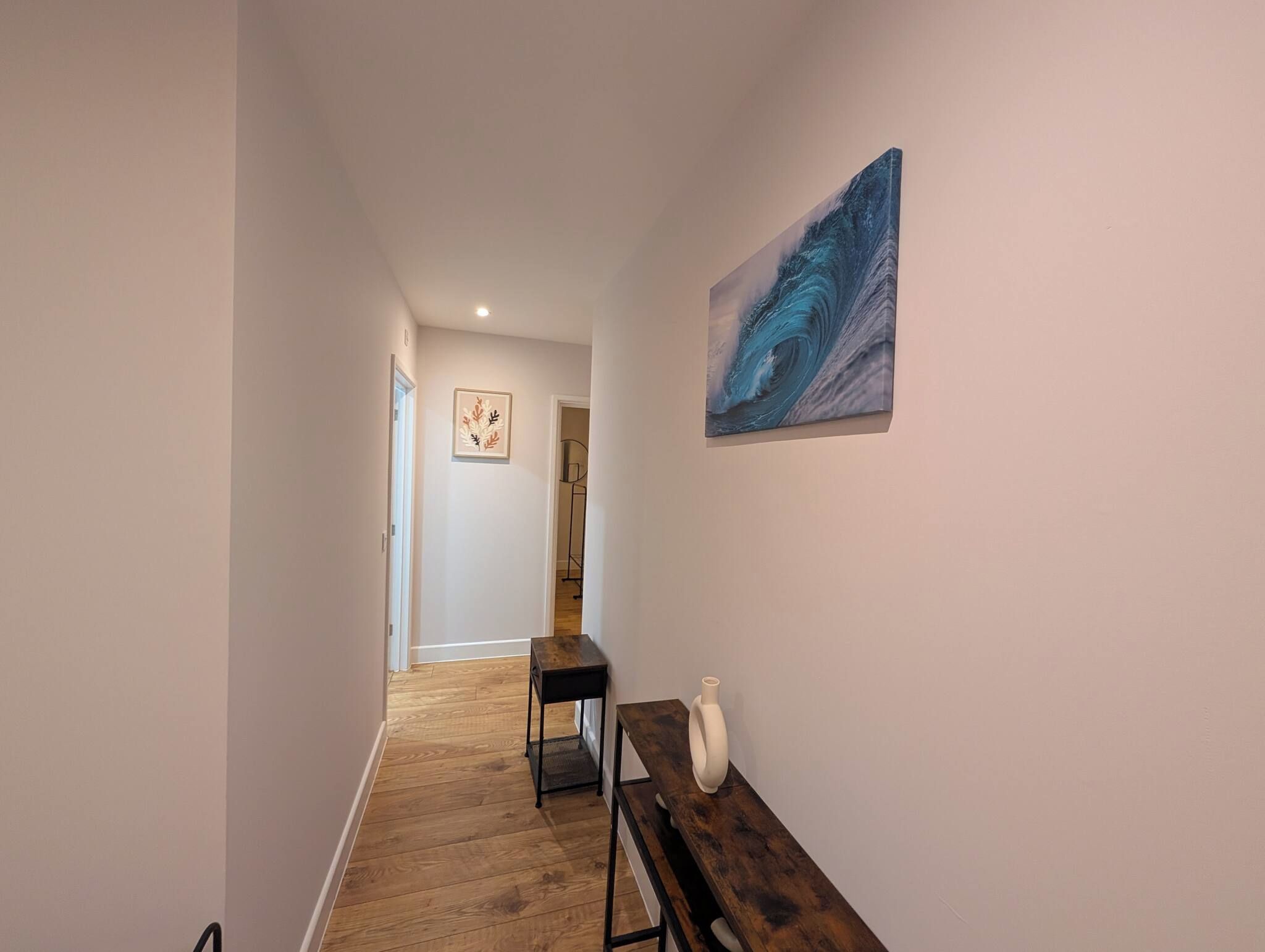 A hallway with wooden flooring, a bench, a white vase, and two artworks on the walls.
