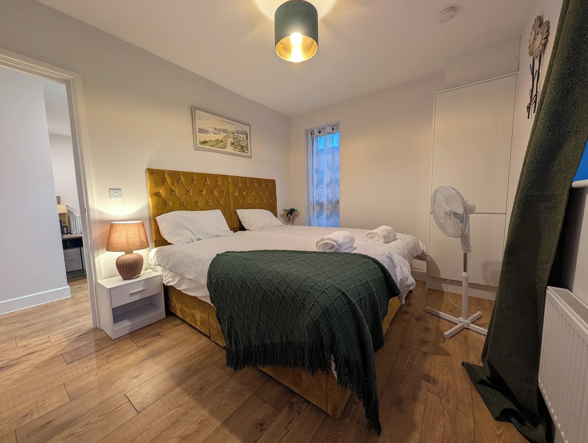 Un dormitorio con cama, mesita de noche, lámpara y ventilador de pie.