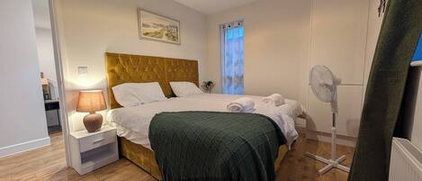 Un dormitorio con cama, mesita de noche, lámpara y ventilador de pie.