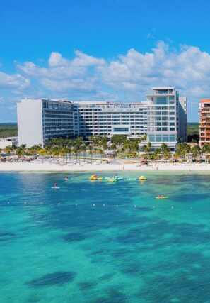 Un hotel frente a la playa con varios pisos, una playa de arena y aguas turquesas cristalinas.