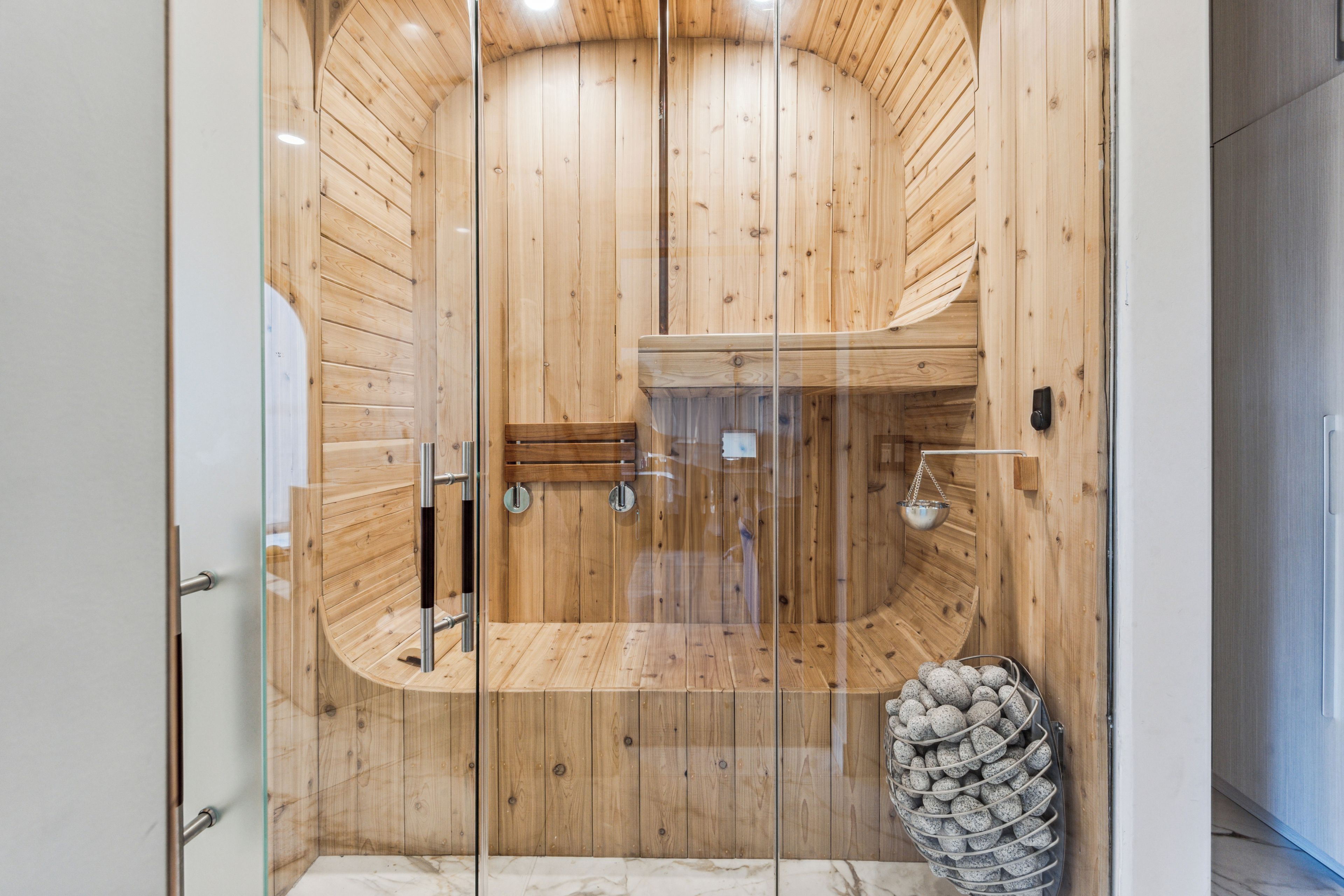 A sauna with wooden walls and a glass door, a bench, and a metal container with stones.