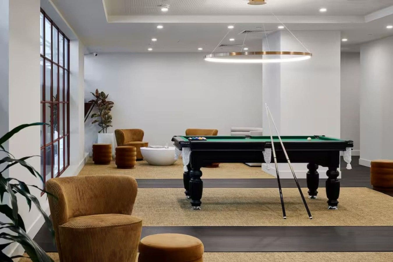 A pool table in a room with a high ceiling, recessed lighting, and a large window.