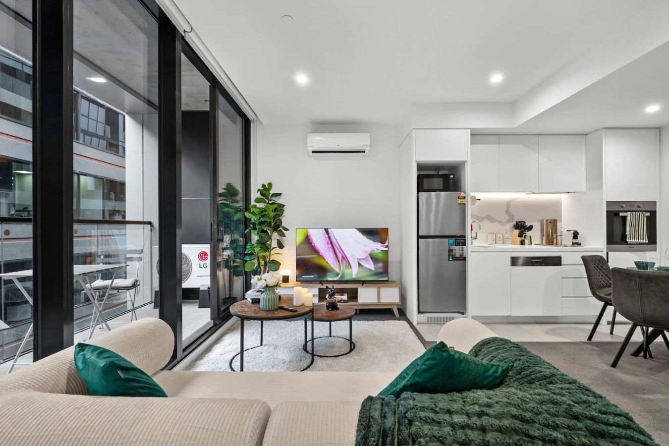 A modern living room with a sofa, a coffee table, and a TV. There is a kitchen area with a fridge and a microwave.