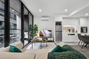 A modern living room with a sofa, a coffee table, and a TV. There is a kitchen area with a fridge and a microwave. - Rooftop Pool Views · Sleek 1BR on St Kilda Rd (Melbourne)