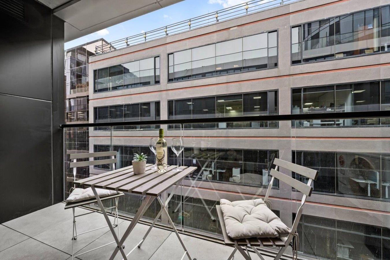 A modern balcony with a glass table, two chairs, a bottle of wine, and a small plant.