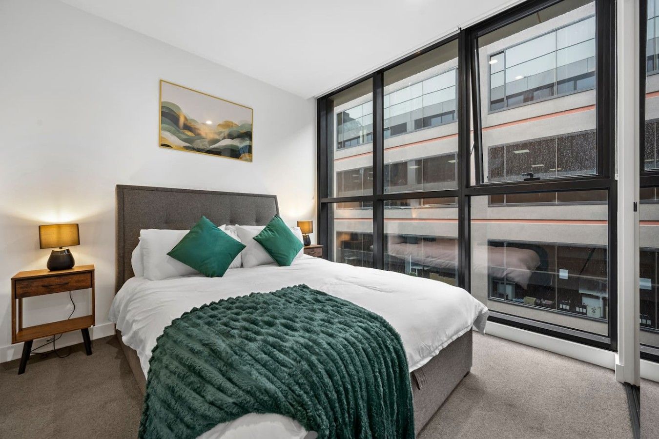 A modern hotel room with a large window, a bed with green pillows, a bedside table with a lamp, and a painting on the wall.