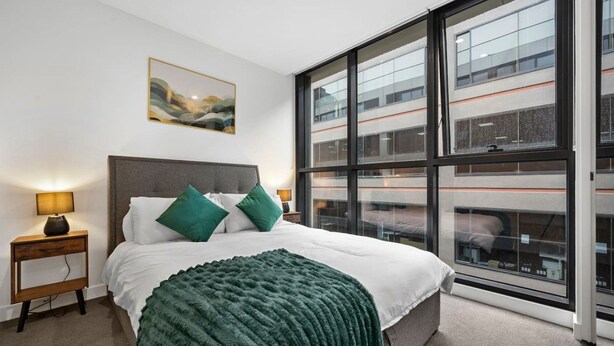 A modern hotel room with a large window, a bed with green pillows, a bedside table with a lamp, and a painting on the wall. - Rooftop Pool Views · Sleek 1BR on St Kilda Rd (Melbourne)