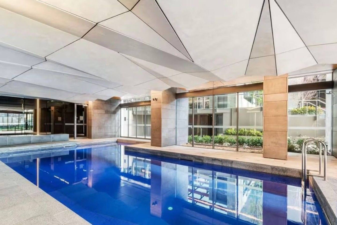 A modern indoor swimming pool with clear glass walls, a tiled ceiling, and a metal ladder.